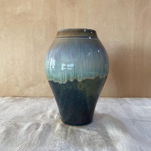 May include: A handcrafted ceramic vase with a glossy finish. The vase features a gradient of colors, transitioning from a dark navy blue base to teal, light blue, and a brown rim. The vase has a rounded body and a slightly flared opening.