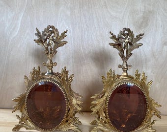 May include: A pair of gold-toned decorative perfume bottles with ornate floral and cherub accents. Each bottle has a round, amber-colored glass insert.