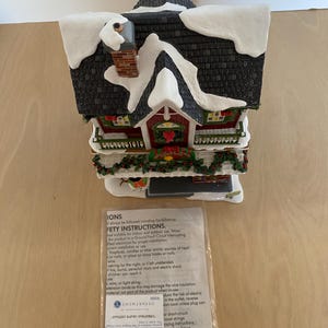 Może przedstawiać: Ceramiczna figurka świątecznego domku z ośnieżonym dachem i kominem. Domek ma czerwone drzwi i okna, z dekoracjami w postaci wieńc&oacute;w i girland. Figurka zawiera małą instrukcję.