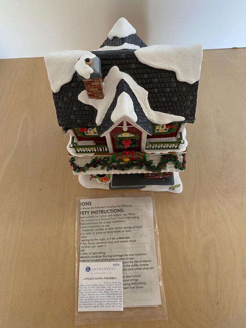 Może przedstawiać: Ceramiczna figurka świątecznego domku z ośnieżonym dachem i kominem. Domek ma czerwone drzwi i okna, z dekoracjami w postaci wieńc&oacute;w i girland. Figurka zawiera małą instrukcję.