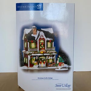 Może przedstawiać: Figurka Department 56 Christmas Crafts Cottage. Dwupiętrowy budynek ma czerwoną ceglaną fasadę, ośnieżony dach i świąteczne dekoracje. Osoba idzie przed domkiem. Widoczny jest tekst "Christmas Crafts Cottage".
