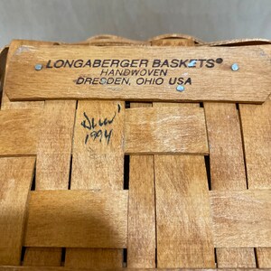 May include: Close-up of a woven wooden basket with a rectangular wooden rim. The rim has the text "LONGABERGER BASKETS HANDWOVEN DRESDEN, OHIO USA". The basket is made of light brown wood and has a signature and the year 1994.