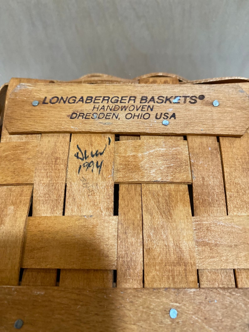 May include: Close-up of a woven wooden basket with a rectangular wooden rim. The rim has the text "LONGABERGER BASKETS HANDWOVEN DRESDEN, OHIO USA". The basket is made of light brown wood and has a signature and the year 1994.