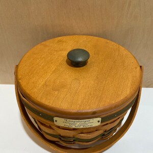 May include: A round wooden basket with a lid and handle. The basket is made of woven wood and has a dark green band around the middle. The lid has a dark knob. A metal tag reads "Longaberger Christmas Collection 1996 Edition Jingle Bell Basket."