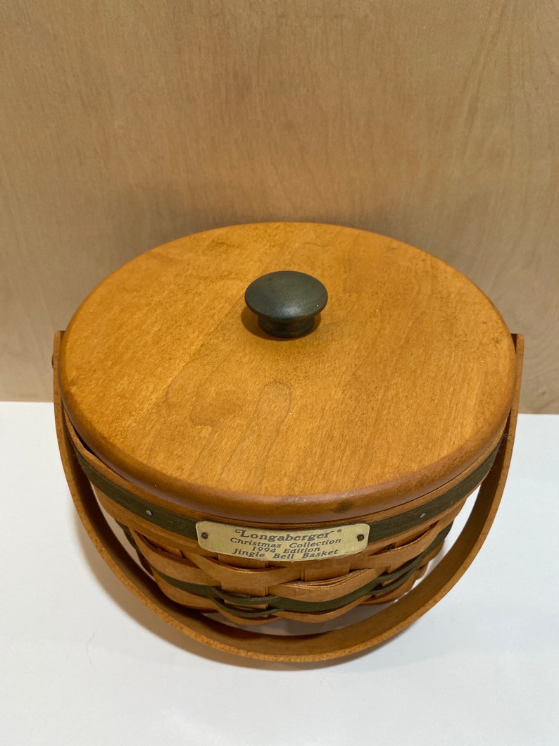 May include: A round wooden basket with a lid and handle. The basket is made of woven wood and has a dark green band around the middle. The lid has a dark knob. A metal tag reads "Longaberger Christmas Collection 1996 Edition Jingle Bell Basket."