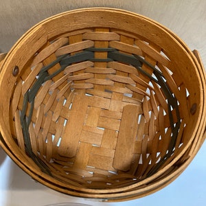 May include: A round woven basket in natural wood tones, with green accents. The basket is made from interwoven wooden strips, creating a textured pattern. It has a handle and a solid base, suitable for storage or display.