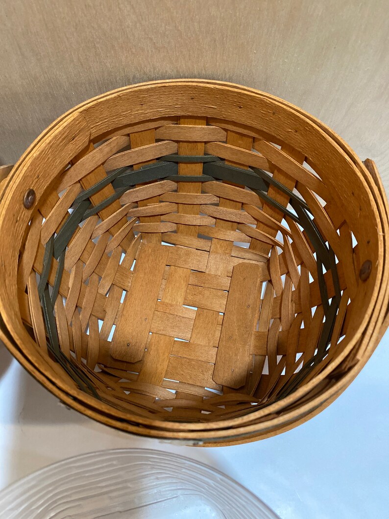 May include: A round woven basket in natural wood tones, with green accents. The basket is made from interwoven wooden strips, creating a textured pattern. It has a handle and a solid base, suitable for storage or display.