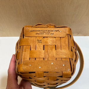 May include: A handwoven Longaberger basket with a handle. The basket is made of light brown woven wood and features a rectangular shape. The text "LONGABERGER BASKETS HANDWOVEN DRESDEN, OHIO USA" is visible on the top. The basket is dated 1994.