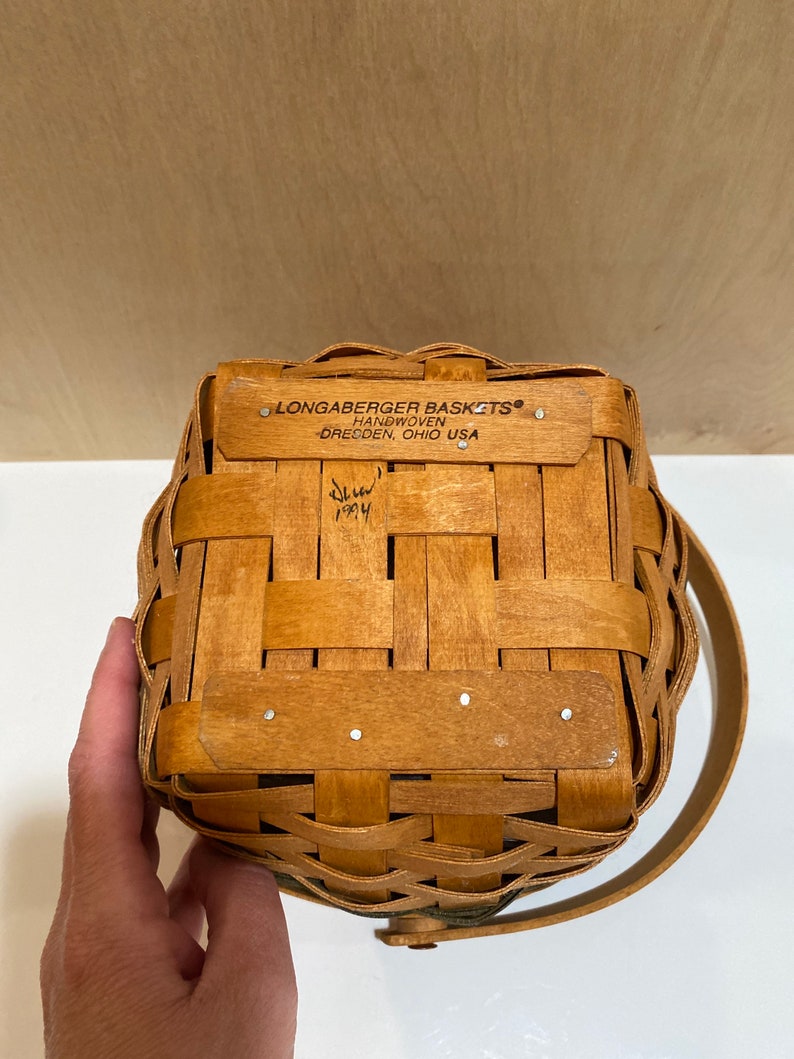 May include: A handwoven Longaberger basket with a handle. The basket is made of light brown woven wood and features a rectangular shape. The text "LONGABERGER BASKETS HANDWOVEN DRESDEN, OHIO USA" is visible on the top. The basket is dated 1994.