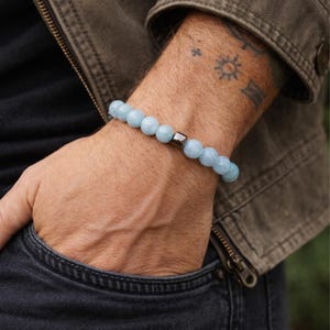 Op de afbeelding: Een lichtblauwe kralenarmband met een donkergrijze metalen accentkraal. De armband is gemaakt van ronde, lichtblauwe kralen. De armband wordt om een pols gedragen, met een donkere jas en jeans op de achtergrond.