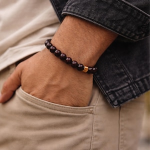 Peut inclure: Un bracelet de perles rouge foncé avec une perle d'espacement dorée. Le bracelet est porté au poignet. On aperçoit une veste en jean et un pantalon beige en arrière-plan. Les perles mesurent environ 0,8 cm de diamètre.