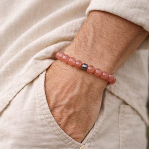 Peut inclure: Bracelet de perles rondes rose-rouge, avec un aspect moucheté. Une seule perle cylindrique, gris foncé, est centrée sur le bracelet. Le bracelet est porté au poignet, avec une chemise de couleur neutre et une poche en arrière-plan.