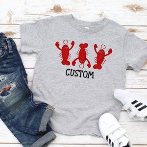 May include: A gray short-sleeved t-shirt with a graphic of three red lobsters with black eyes and the word "CUSTOM" printed below. The shirt is paired with blue jeans and white sneakers with black stripes.