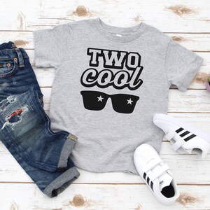 May include: A gray short-sleeved t-shirt with the text "TWO cool" in black with sunglasses and stars printed on it.