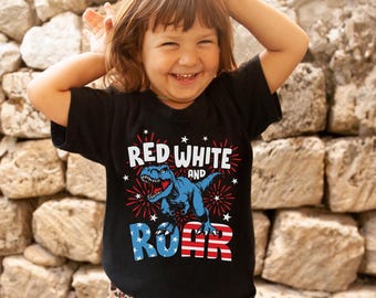 May include: A black t-shirt with the text "RED WHITE AND ROAR" and a blue dinosaur graphic. The text and dinosaur are in red, white, and blue colours. The child is wearing leopard print trousers.