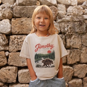 May include: A beige short-sleeve t-shirt featuring a graphic print of a forest landscape with a bear, a red sun, and the text Jimothy Feral Legend in red script. The shirt has a crew neckline and a relaxed fit, showcasing a nature-inspired design.