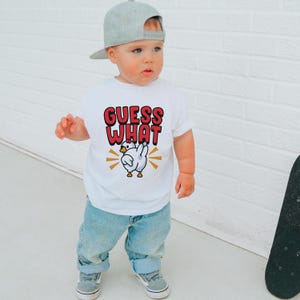 May include: A white t-shirt with the text "GUESS WHAT" in red and black, featuring a cartoon chicken graphic. The child is wearing light blue jeans, gray shoes, and a gray baseball cap.