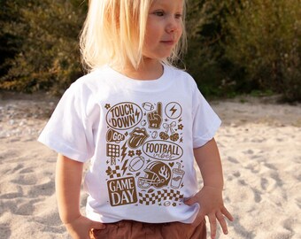May include: A white t-shirt featuring a brown graphic design with football-themed illustrations and text. The design includes a football helmet, a football, and the words "TOUCH DOWN", "GAME DAY", and "FOOTBALL vibes".