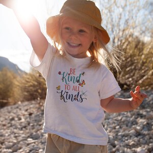 May include: A white t-shirt with the words "BE Kind TO ALL Kinds" in a colorful design with butterflies, bees, and a ladybug. The child is wearing a yellow hat.