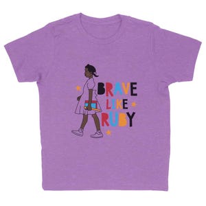 May include: A lilac-colored t-shirt featuring a graphic of a young girl walking, carrying books. The text "BRAVE LIKE RUBY" is printed in colorful letters. The design includes stars, creating a cheerful and inspiring look.