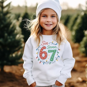 May include: A white sweatshirt with the text "Six Seven" and the numbers "67" in red and green, decorated with colorful lights. The sweatshirt is worn by a person wearing a white beanie.