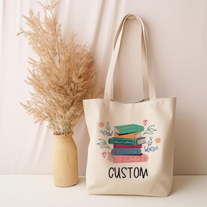 May include: A beige canvas tote bag with a stack of colorful books and flowers printed on it. The text "CUSTOM" is printed below the books.