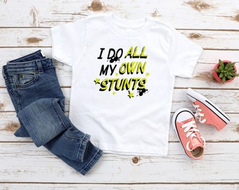 May include: White t-shirt with the text "I do all my own stunts" in yellow and black lettering with yellow stars. The shirt is paired with blue jeans and pink sneakers.