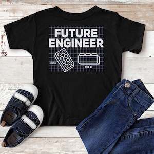 May include: Black t-shirt with white text that reads "FUTURE ENGINEER" and a diagram of two building blocks. The shirt is laid out on a wood surface with a pair of blue jeans and blue sneakers.