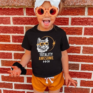 May include: A child wearing a black t-shirt with a cat wearing eclipse glasses and the text "Totality Awesome 4-8-24". The child is also wearing a white headband and orange sunglasses.