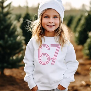 May include: A young girl wearing a white sweatshirt and a white beanie smiles at the camera. The sweatshirt features the number "67" in pink, with the words "SIX" and "SEVEN" below the numbers.