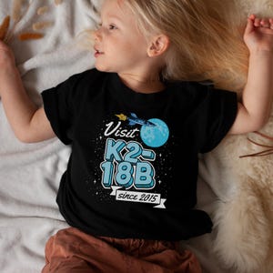 May include: Black t-shirt with a blue and white graphic of a rocket ship flying past a planet with the text "Visit K2-18B since 2015".