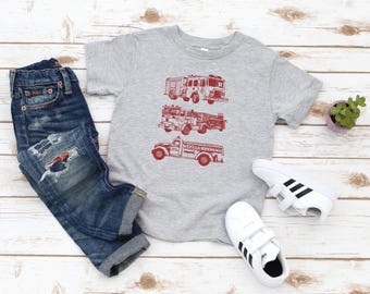 Puede incluir: Una camiseta gris con un gráfico rojo de tres camiones de bomberos. La camiseta se combina con jeans azules y zapatillas blancas.