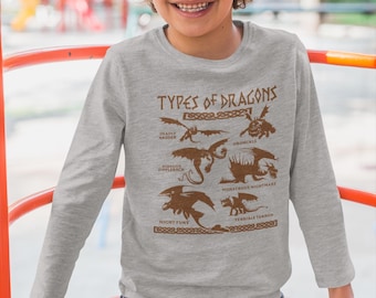 May include: A gray long-sleeve shirt featuring a brown graphic of different dragon types with the text "TYPES OF DRAGONS". The shirt is worn by a child wearing a blue beanie and blue jeans.