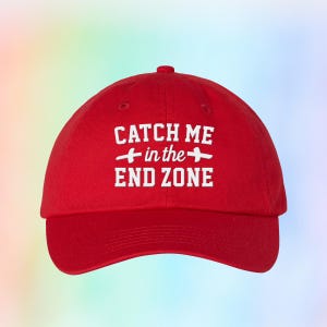 May include: A red baseball cap with the text "CATCH ME in the END ZONE" in white. The cap has a curved brim and a rounded crown. The text is centered on the front of the cap.