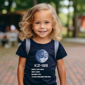 May include: A young child wearing a navy blue t-shirt featuring a graphic of the planet K2-18B. The shirt includes the planet's radius (8,867.6 miles), orbit (33 days), discovery year (2015), and distance from Earth (124 light-years).