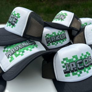 May include: A stack of black and white trucker hats with green pixelated text. The text on the hats reads "Maverick", "Jacob", "Makar", and "Sage".