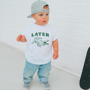 May include: A toddler wearing a white t-shirt with the word "LATER" above a green alligator graphic. The child is also wearing light blue jeans, gray sneakers, and a gray baseball cap.