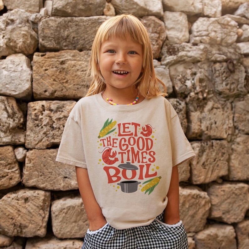 May include: A child wearing a beige t-shirt with a red, yellow, and black graphic that says "Let the good times boil". The graphic includes a pot, corn, and shrimp. The child is standing in front of a stone wall.