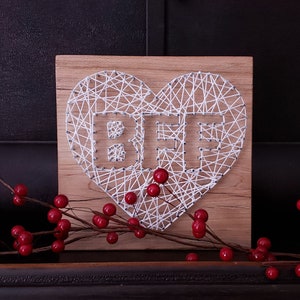 May include: A wooden heart-shaped sign with the word "BFF" spelled out in white string. The sign is on a dark surface with red berries.