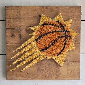 Op de afbeelding: Een string art-stuk dat het logo van de Phoenix Suns weergeeft. Het logo is een gestileerde zon met een basketbal in het midden. De zon is gemaakt van gele en oranje draad, en de basketbal is gemaakt van oranje en zwarte draad.
