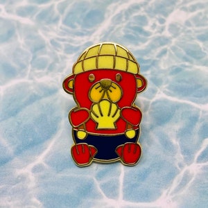 May include: A colorful enamel pin featuring a cartoon bear character. The bear is primarily red with yellow and blue accents, wearing a yellow hat and holding a yellow seashell. The pin has a gold-colored outline.