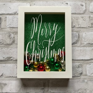 Könnte beinhalten: Ein weiß gerahmter Weihnachtsschattenkasten mit grünem Glitzerhintergrund. Die Worte "Merry Christmas" sind in weißer Schrift geschrieben. Die Box ist mit roten, grünen, goldenen und silbernen Weihnachtskugeln gefüllt.