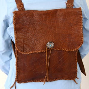 Puede incluir: Un bolso de cuero marrón con cierre de solapa y un botón de metal decorativo. El bolso tiene un borde con flecos y se lleva sobre el hombro.