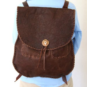 Puede incluir: Una mochila de cuero marrón con cierre de solapa y un botón de hueso decorativo. La mochila tiene dos bolsillos en la parte delantera y está hecha con un diseño cosido a mano.