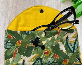 May include: A yellow fabric glasses case with a tropical print featuring toucans, parrots, and palm leaves. The case is open and a pair of black cat-eye glasses are inside.