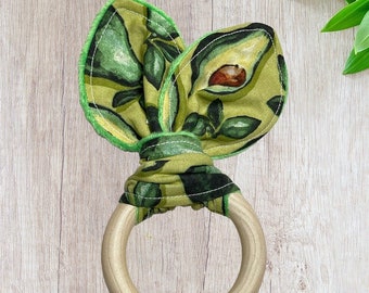 May include: A wooden teething ring with a green and yellow fabric bow tied around it. The fabric has a pattern of avocados.