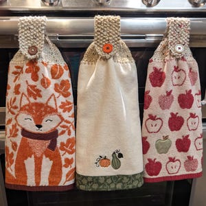 May include: Three hanging kitchen towels with knit tops and buttons. The left towel features an orange fox and autumn leaves. The centre towel is cream with a small pumpkin and gourd embroidery. The right towel displays a red and green apple pattern.