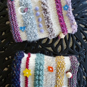 May include: Two handmade sensory pads with various textures and embellishments. Each pad features knitted sections in cream, gray, and colorful stripes. Adorned with buttons, beads, and soft textures, these pads offer tactile exploration for sensory development.
