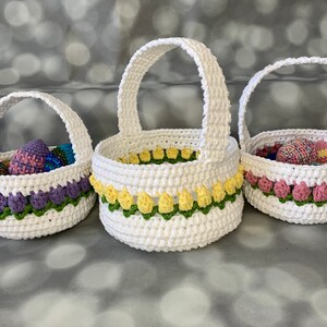 以下が含まれることがあります： 異なるサイズで、それぞれ異なる色の花トリムが付いた、3つのクロシェ編みイースターバスケット。最も小さいバスケットは、紫、ピンク、青のイースターエッグでいっぱいです。真ん中のバスケットは、黄色と緑の花トリムが付いています。最も大きいバスケットは、ピンクと緑の花トリムが付いています。