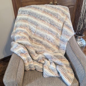 May include: A cozy, hand-knitted blanket draped over a chair. The blanket features a striped pattern in shades of cream, gray, and peach. The chair is upholstered in a neutral gray fabric, creating a warm and inviting scene.